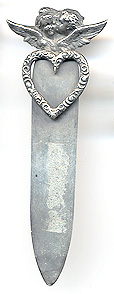 This bookmark was made in the US by an unknown manufacturer. It is marked only sterling. It depicts two cherub heads, a girl and a boy where the boy is kissing the girl. The heads are above two wings. The top blade is a cut out heart shape. The date is 1900 - 1910.